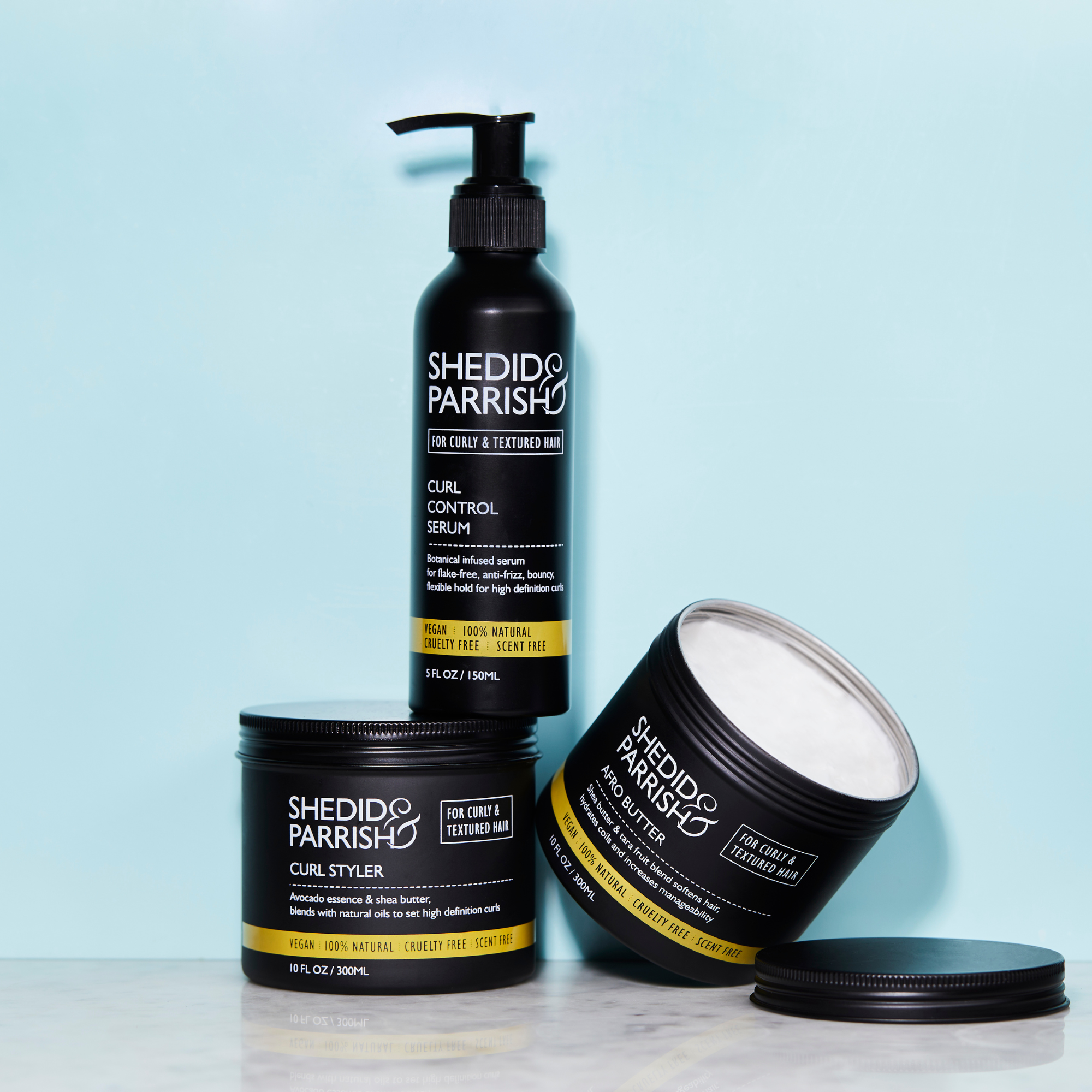 Trio of Shedid & Parrish styling products in aluminium packaging with the afro butter lid removed.  Sat on a marble table with a light blue background